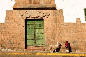 Jauna mergina ir jos alpaka, einančios palei rūmų sieną, dekoruotą inku simboliais. Peru, Cuzco regionas,, Cuzco, Nezaneras aikštė 